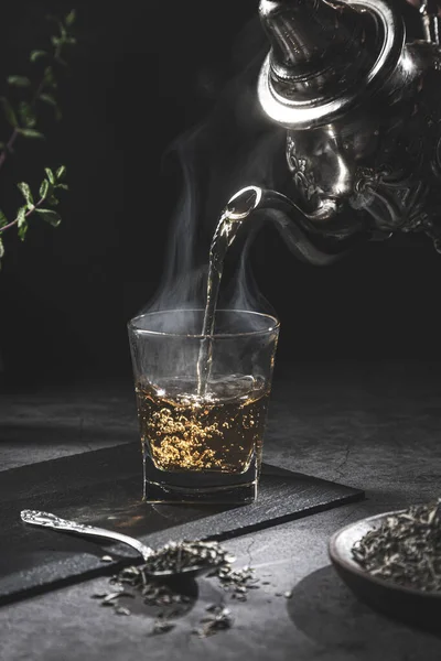 Traditional Moroccan teapot pouring tea into a steaming glass with natural tea and mint, on a dark background. Traditional Moroccan tea concept.