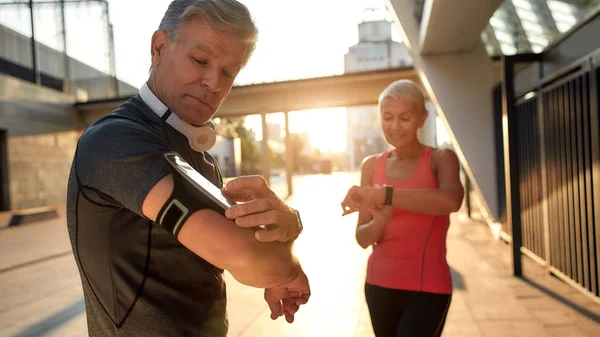 Używając nowoczesnych technik. Aktywna para w średnim wieku w odzieży sportowej sprawdza wyniki treningu stojąc razem na świeżym powietrzu. Krokomierz kontrolny — Zdjęcie stockowe