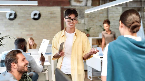 El poder del trabajo en equipo. Joven hombre asiático positivo en ropa casual sosteniendo tableta digital y explicando algo a sus colegas mientras trabajan juntos en la oficina moderna — Foto de Stock