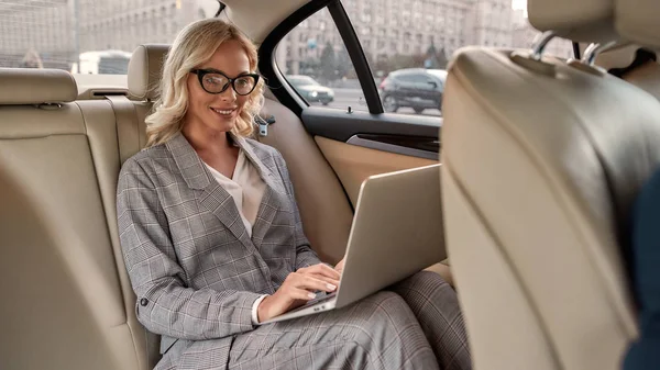Szczęśliwa bizneswoman. Atrakcyjna bizneswoman w stylowym klasycznym ubraniu pracująca na laptopie i uśmiechnięta siedząc w samochodzie — Zdjęcie stockowe