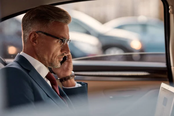 Busy businessman. Side view of mature man in classic wear sitting in the car and talking by the phone with client