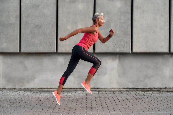 Full of energy. Side view of active middle aged woman in sport clothing jumping while exercising outdoors