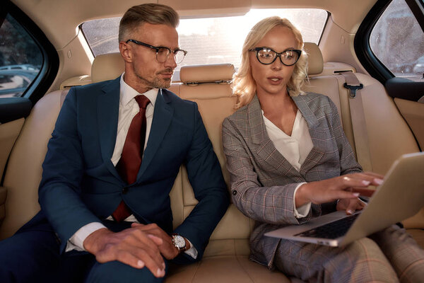 Look here. Young and beautiful business woman in classic wear showing something on laptop to her business partner while sitting together in the car