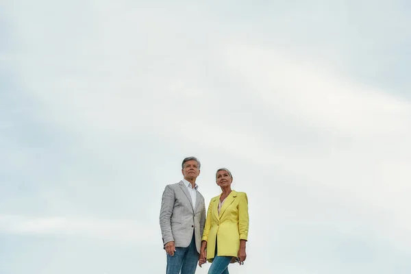 Sólo felices juntos. Elegante y encantadora pareja de mediana edad cogida de la mano y mirando hacia otro lado mientras está de pie contra el cielo azul —  Fotos de Stock