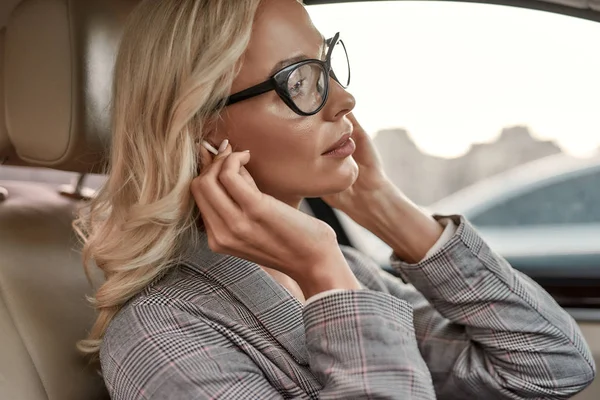 Η αγαπημένη μου λίστα αναπαραγωγής. Ελκυστική και στιλάτη επιχειρηματίας με γυαλιά ηλίου που βάζει ακουστικά στα αυτιά της ενώ κάθεται στο κάθισμα του οδηγού στο αυτοκίνητο — Φωτογραφία Αρχείου