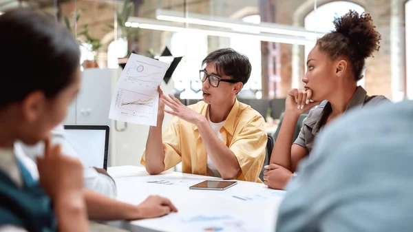 Analisando resultados. Jovem asiático apontando para relatório financeiro e explicando algo para seus jovens colegas enquanto sentado no escritório moderno — Fotografia de Stock