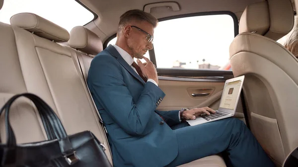 Viagem de negócios. Vista lateral de belo homem de negócios maduro em terno completo trabalhando em seu laptop enquanto sentado no carro — Fotografia de Stock