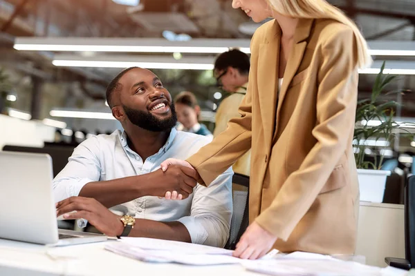 Lo logramos. Joven hombre alegre afro americano alegre y hermosa mujer de negocios estrechando la mano mientras trabajan juntos en la oficina moderna — Foto de Stock