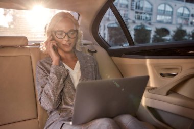 Proje detaylarını tartışıyoruz. Kendine güvenen, takım elbiseli bir iş kadını dizüstü bilgisayarla çalışıyor ve arabada iş ortağıyla telefonda konuşuyor..