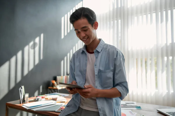 Απολαμβάνει τη δουλειά του. Πορτρέτο του νεαρού χαρούμενου Ασιάτη που χρησιμοποιεί το touchpad και χαμογελά ενώ ακουμπά σε ένα τραπέζι στο σύγχρονο γραφείο. Ευτυχής σχεδιαστής που εργάζεται με ψηφιακή ταμπλέτα — Φωτογραφία Αρχείου
