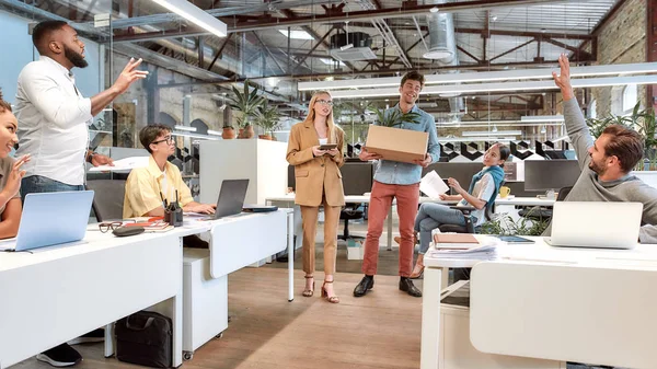Neues Teammitglied. junger Mann in Freizeitkleidung hält Schachtel mit Dingen, während ihn seine neuen Mitarbeiter im modernen Büro begrüßen — Stockfoto