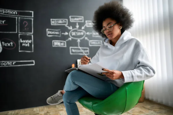 Olvasok a munkahelyemen. Fiatal afro-amerikai női grafikus szemüveg olvasás magazin, miközben ül a széken ellen táblán a nagy kreatív irodában. Kreatív ügynökség — Stock Fotó