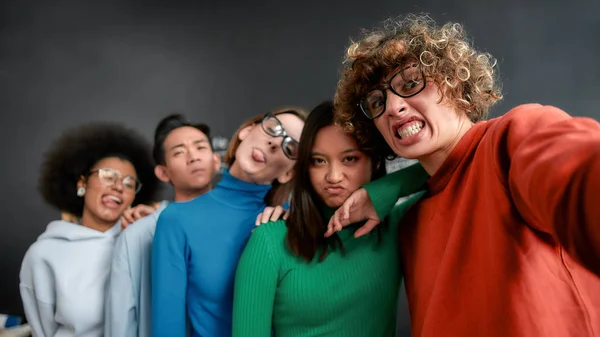 Funny selfie. Positive group of multicultural people in casual wear making grimaces and looking at camera while making a selfie against blackboard in the creative office — Stock Photo, Image