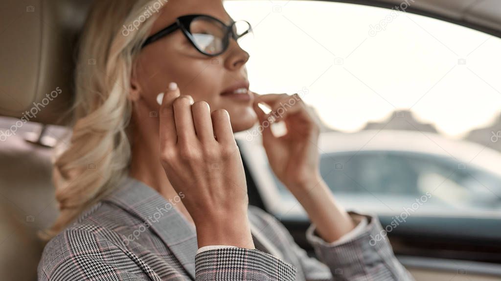 Listening audiobook. Attractive and stylish businesswoman in eyeglasses putting headphones into her ears while sitting on drivers seat in the car