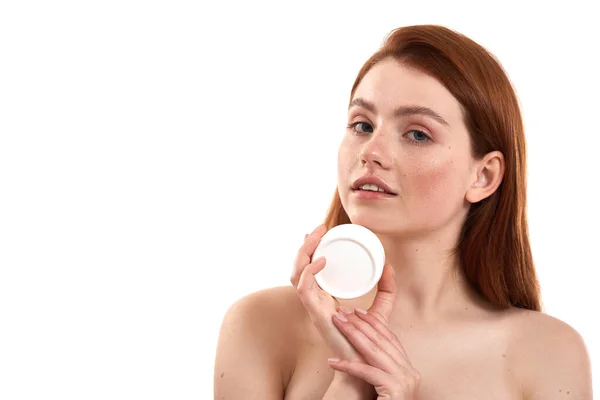 Taking care of her sensitive skin. Young attractive red-haired girl with freckles demonstrating cosmetic product and looking at camera while standing in studio against white background — Stock Photo, Image