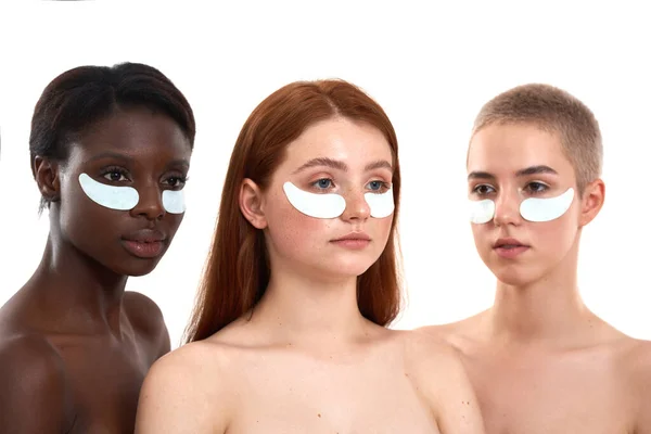 Když se probudíš s temnými kruhy. Tři nádherné multikulturní mladé ženy, které používají pásky přes oko, zatímco stojí těsně vedle sebe na bílém pozadí. Různé etnické ženy — Stock fotografie