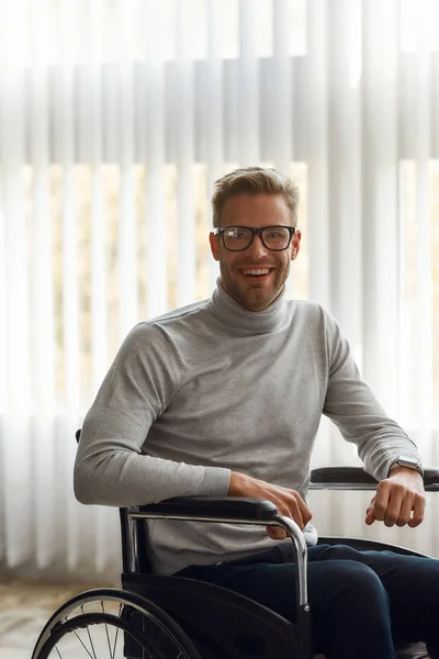 Great mood. Portrait of handsome cheerful male office worker in a wheelchair in glasses looking at camera and smiling while sitting at his workplace in modern office — Stock Photo, Image