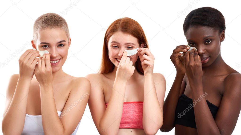Spa girls. Three gorgeous multicultural young women applying eye patches under the eyes and smiling while standing against white background. Different ethnicity women