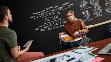 Tekerlekli sandalyedeki genç Ux Ui tasarımcısı elinde kitap tutarken ve yaratıcı ofisteki sunumuyla tahtada otururken meslektaşıyla bazı fikirleri tartışıyor.