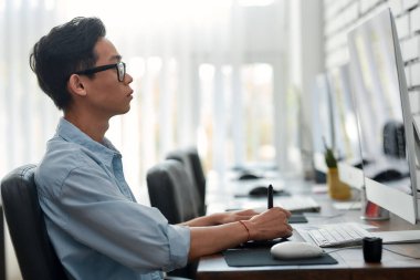 Yaratıcı olmak. Dijital grafik tableti kullanan gözlüklü Asyalı genç bir grafik tasarımcının yaratıcı ajansındaki iş yerinde otururken yan görüntüsü