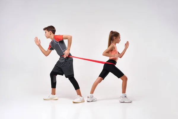 Fitness leaders. Full-length shot of teenage boy and girl engaged in sport, looking focused while exercising using resistance band. Isolated on white background. Training, active lifestyle concept — Stock Photo, Image