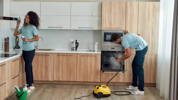 Garder votre maison propre. Deux jeunes nettoyeurs professionnels en uniforme travaillent ensemble dans la cuisine. Jeune homme caucasien utilisant un nettoyeur à vapeur, femme afro américaine heureuse nettoyage hotte de cuisine . — Photo