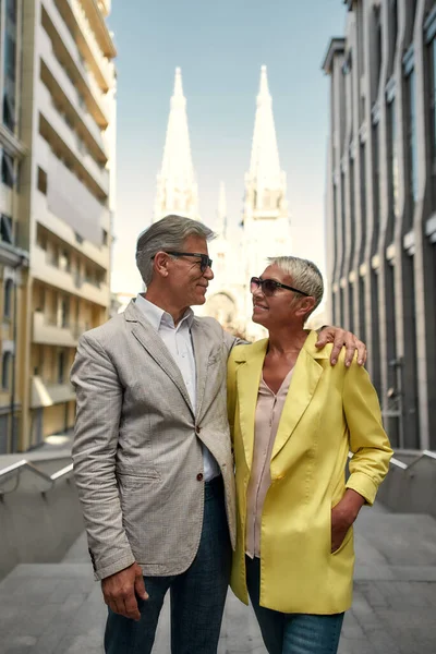 Modern couple. Elegant and stylish mature couple in sunglasses hugging and looking at each other with smile while standing outdoors against beautiful catholic church