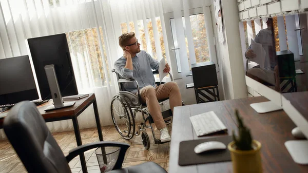 Op zoek naar inspiratie. Portret van jonge geconcentreerde mannelijke kantoormedewerker in een rolstoel die documenten op wit bord voor zich bekijkt in het heldere moderne kantoor — Stockfoto