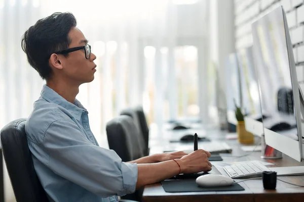 Ser creativo. Vista lateral de un joven diseñador gráfico asiático en gafas usando una tableta gráfica digital mientras está sentado en su lugar de trabajo en la agencia creativa —  Fotos de Stock