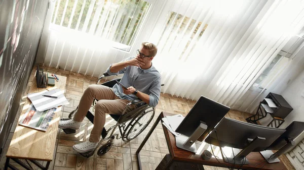 Thinking about business. Thoughtful young male office worker in a wheelchair holding digital tablet and analyzing at charts and graphs on blackboard while working in the modern office — Stock Photo, Image