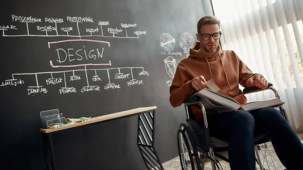 Konzentriert auf das Lesen. Porträt eines jungen professionellen Designers im Rollstuhl, der ein Buch liest, während er neben der Tafel sitzt und im Kreativbüro präsentiert wird — Stockfoto