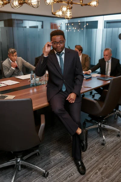 Zakelijk professioneel. Portret van een Afrikaanse zakenman in formele kleding die zijn bril aanpast en naar de camera kijkt terwijl hij naar het bureau leunt — Stockfoto