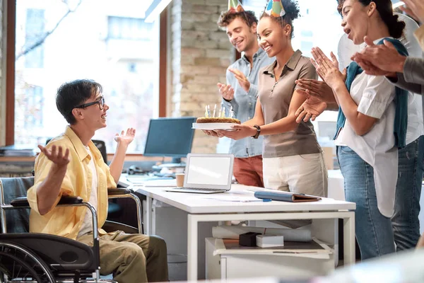 令人惊讶。一群积极的商界人士在现代办公室里庆祝他们的年轻同事坐着轮椅过生日。生日蛋糕 — 图库照片