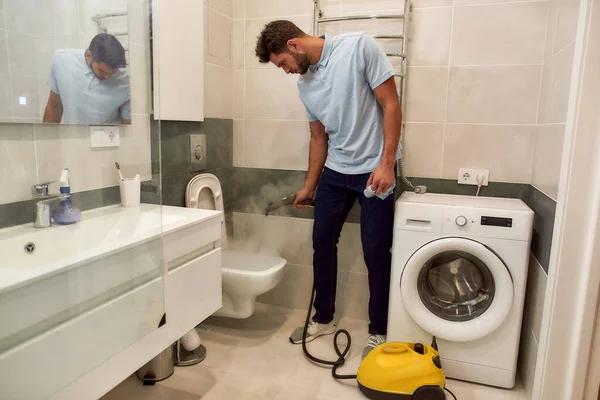 Zawodowiec w pracy. Młody biały mężczyzna w mundurze czyści toaletę z odkurzaczem parowym. Mężczyzna sprzątaczka pracuje w łazience — Zdjęcie stockowe