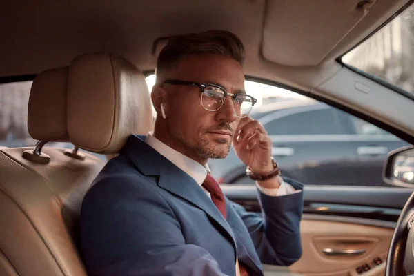 Ik werk overal vandaan. Knappe volwassen man in volle pak aanpassen van de hoofdtelefoon tijdens het besturen van een auto — Stockfoto