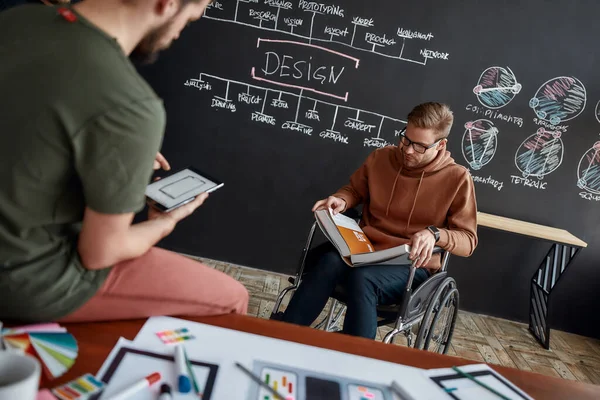 Op zoek naar inspiratie. Geconcentreerde mannelijke Ux Ui ontwerper in een rolstoel die een boek leest terwijl hij bij het schoolbord zit met presentatie in het creatieve kantoor — Stockfoto