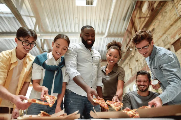 Después del trabajo duro. Un equipo multicultural positivo comiendo pizza y sonriendo mientras almuerza en la oficina moderna — Foto de Stock