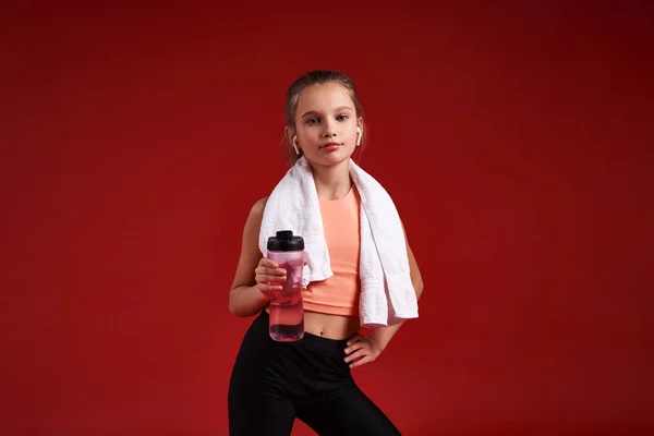 Descansando. Una linda chica dedicada al deporte, ella está mirando a la cámara mientras está de pie con una toalla y una botella de agua. Aislado sobre fondo rojo. Fitness, entrenamiento, concepto de estilo de vida activo. Disparo horizontal — Foto de Stock