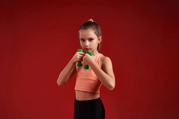 Seien Sie vorbereitet. Ein nettes Kind, Mädchen treibt Sport, sie schaut in die Kamera, während sie mit Hanteln trainiert. Vereinzelt auf rotem Hintergrund. Fitness, Training, aktiver Lebensstil. Horizontalschuss — Stockfoto