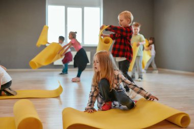 Jimnastik dersi. Bir grup hoş çocuk, günlük kıyafetler içinde sarı minderleri yere seriyor ve dans stüdyosunda yoga dersine hazırlanıyor.