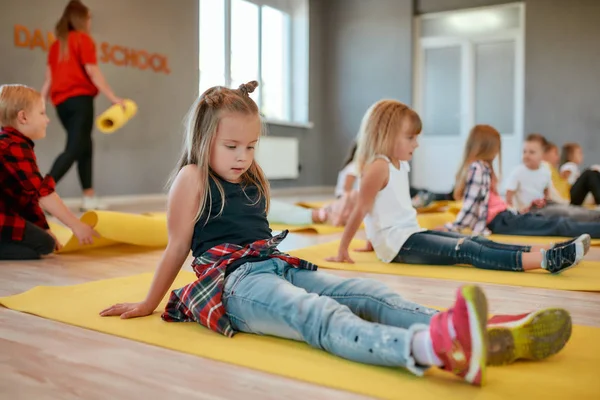 Πορτρέτο ενός όμορφου μικρού κοριτσιού που κάθεται στο κίτρινο στρώμα γιόγκα ενώ έχει μάθημα γιόγκα στην σχολή χορού. Παιδιά που κάνουν γυμναστικές ασκήσεις — Φωτογραφία Αρχείου