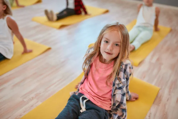 Niños y deporte. Retrato de una niña bonita sentada en la esterilla de yoga amarilla y mirando a la cámara mientras tiene una clase de gimnasia en el estudio de baile — Foto de Stock