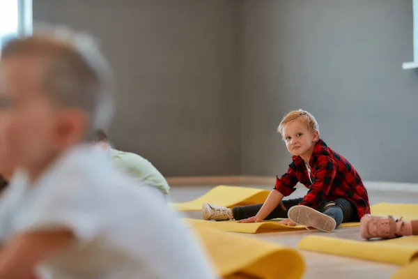 Humor alegre. Retrato de um menino engraçado sentado no tapete de ioga, esticando e olhando para a câmera enquanto se aquece no estúdio de dança — Fotografia de Stock