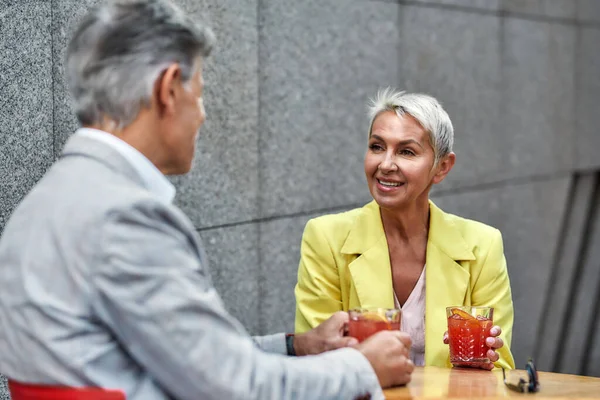 Pasando un buen rato juntos. Mujer alegre y hermosa de mediana edad en traje amarillo chaqueta beber cócteles con su amigo mientras se sienta en la cafetería al aire libre juntos —  Fotos de Stock