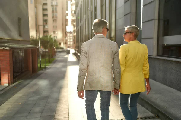Achteraanzicht van stijlvol echtpaar van middelbare leeftijd dat elkaars hand vasthoudt en elkaar aankijkt terwijl ze samen door de straat lopen — Stockfoto