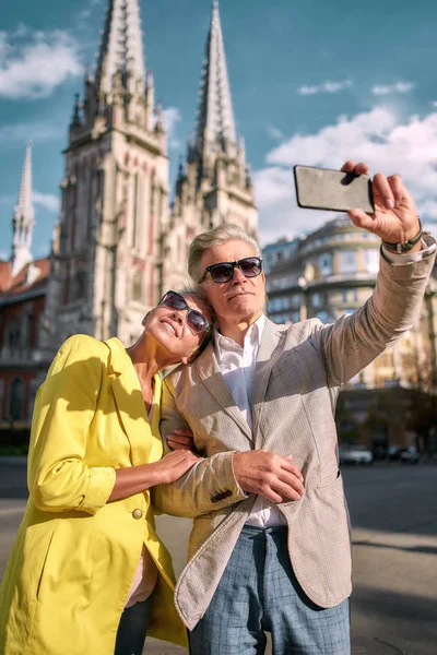 Traveling together. Happy middle-aged caucasian couple in sunglasses taking selfie by mobile phone and smiling while standing against beautiful catholic church