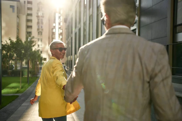 Quero mostrar-te uma coisa. Mulher caucasiana de meia-idade alegre e elegante em óculos de sol segurando a mão dos homens e levando-o a andar ao ar livre — Fotografia de Stock