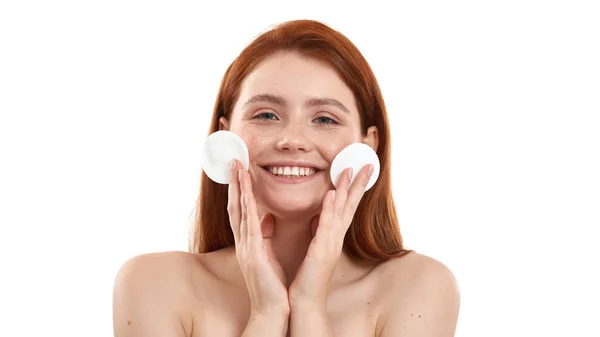 Daily routine. Portrait of happy half naked woman with ginger hair removing makeup from face with cotton pads and smiling while standing in studio against white background — Stock Photo, Image