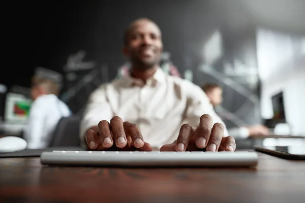 Guida un futuro migliore. Commerciante africano focalizzato seduto alla scrivania e che studia i rapporti analitici utilizzando il pc in ufficio. I suoi colleghi stanno lavorando sullo sfondo. Focus su dita e tastiera — Foto Stock