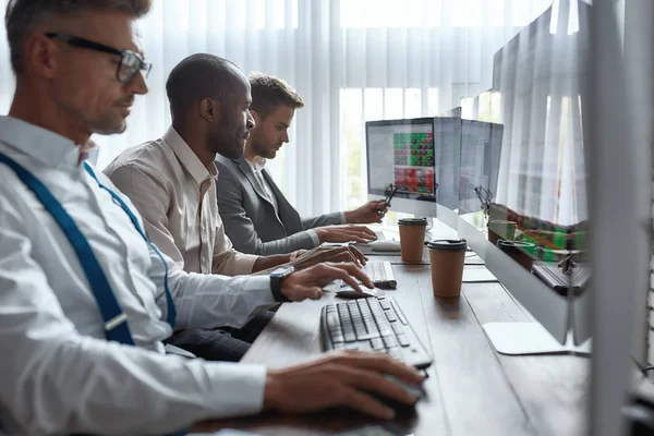 Try and try, earn and learn. Three businessmen sitting at desktop front PC computers with financial graphs and statistics on monitors. Analysis of digital market and investment. Stock trade — Zdjęcie stockowe